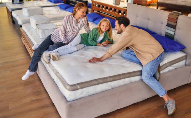 A young family with their lovely daughter lay down on the bed, trying the mattress for softness in the store. Buying a bed and bedding.